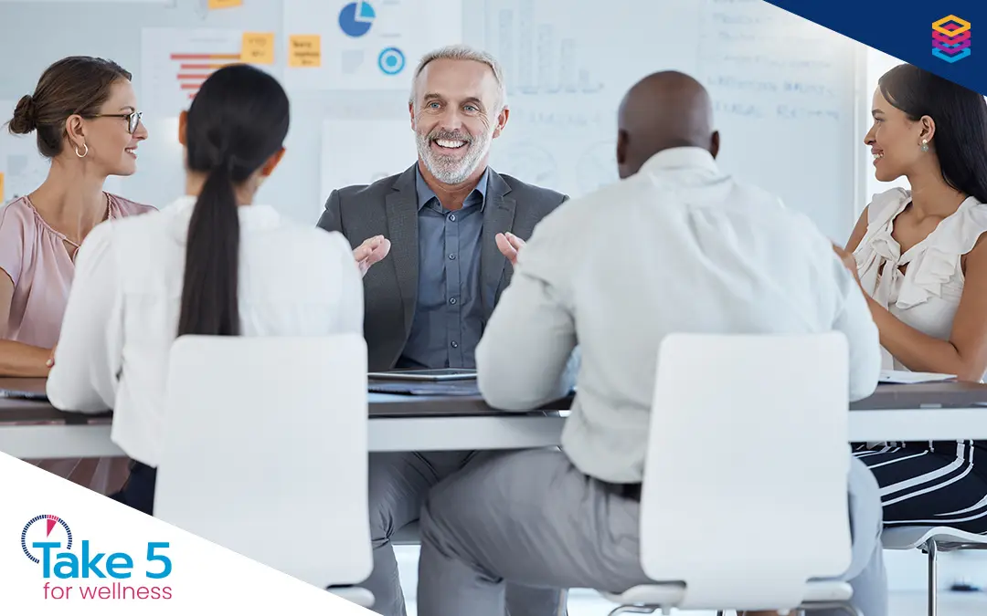 Training unlocks improved mental health | Older man talking to a group in a meeting room while smiling - Benefits Alliance