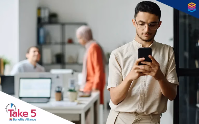 Modern-Day Addictions | Young office worker checking his phone away from his coworkers - Benefits Alliance
