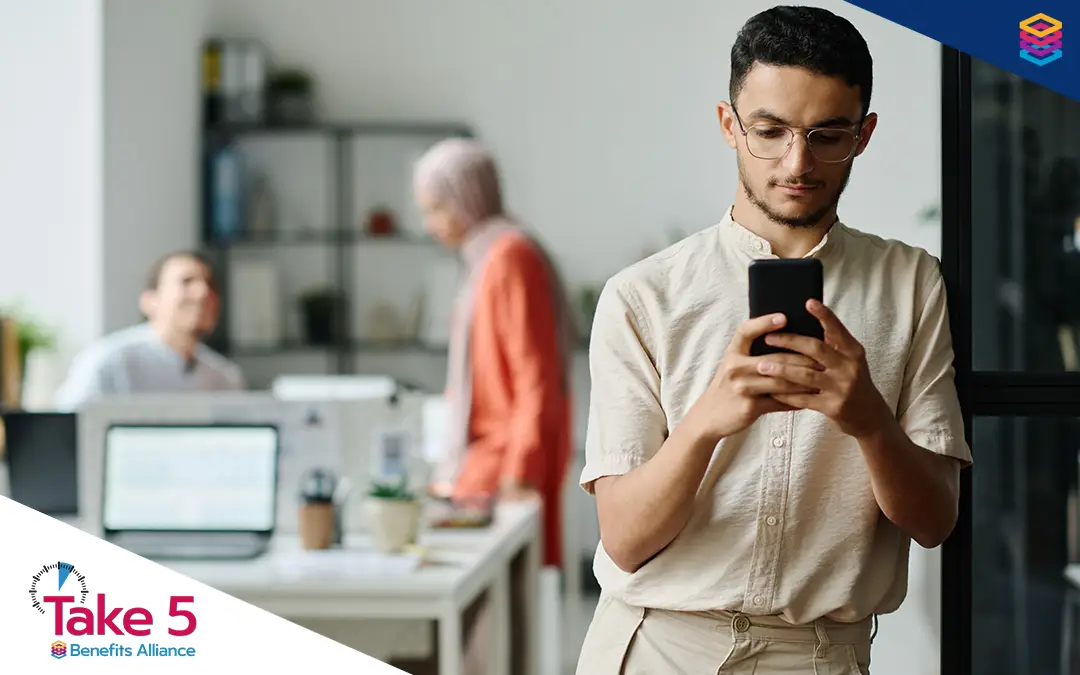 Modern-Day Addictions | Young office worker checking his phone away from his coworkers - Benefits Alliance